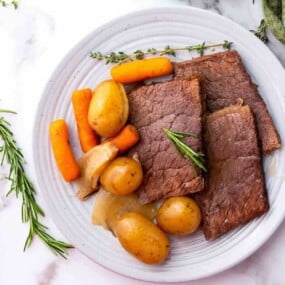 A white plate with slices of cooked beef, baby potatoes, carrots, onions, and garnished with rosemary and thyme.