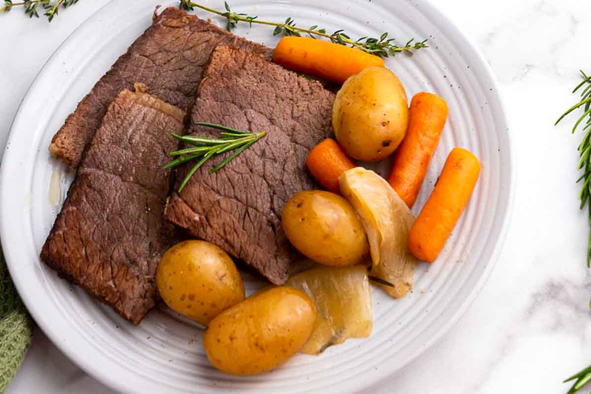 A plate with slices of cooked beef, baby potatoes, carrots, onion pieces, and a sprig of rosemary on a white background.