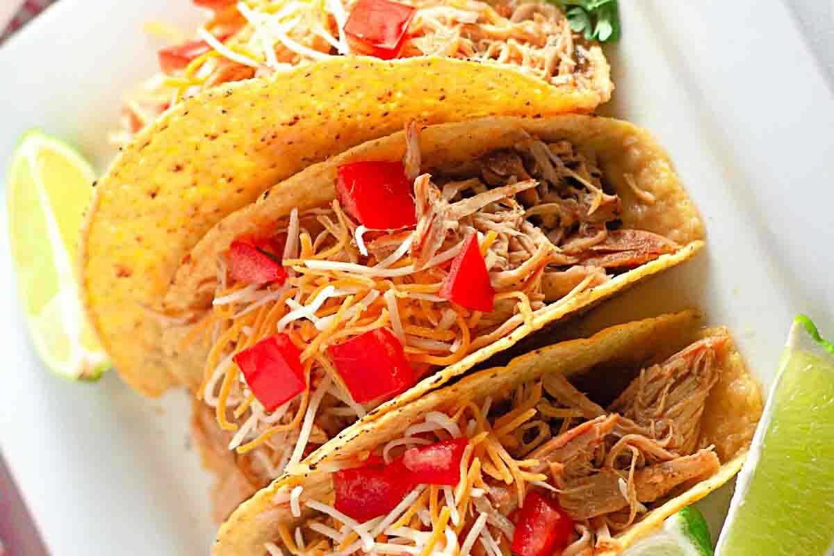 Three hard-shell tacos filled with shredded meat, shredded cheese, and diced tomatoes are served with lime wedges on a white plate.