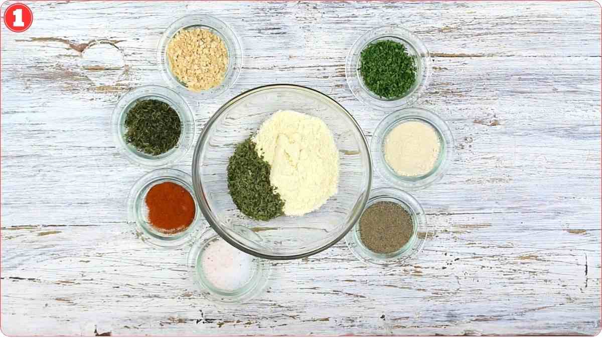 Eight small glass bowls containing various dried herbs and spices arranged in a circle on a white wooden surface.