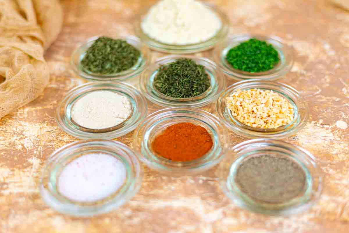 Nine small glass bowls with various dried spices and herbs arranged on a textured brown surface.