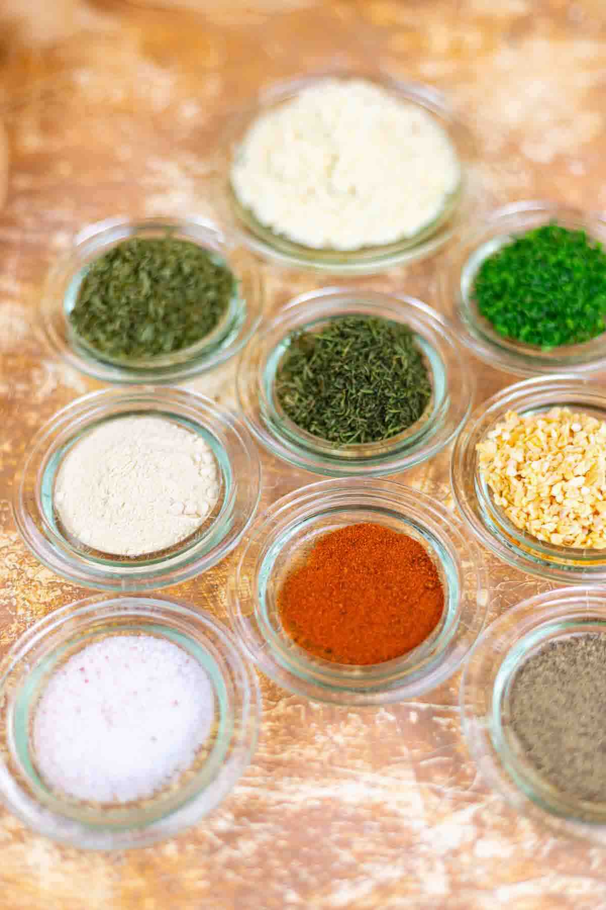 Nine small glass bowls containing various dried herbs and spices, arranged on a light brown textured surface.