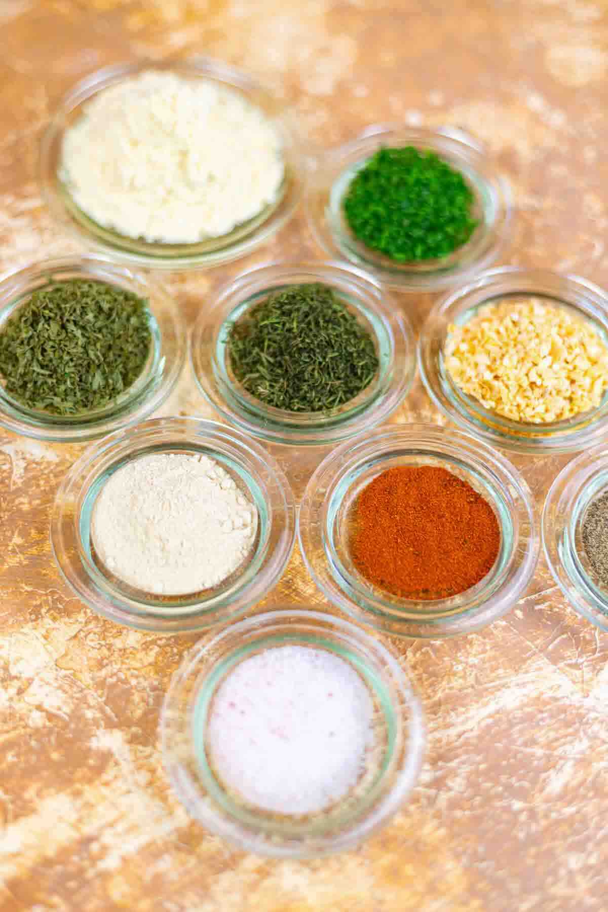 Nine small glass bowls containing various dried herbs and spices, including powders and flakes, are arranged on a textured, light brown surface.
