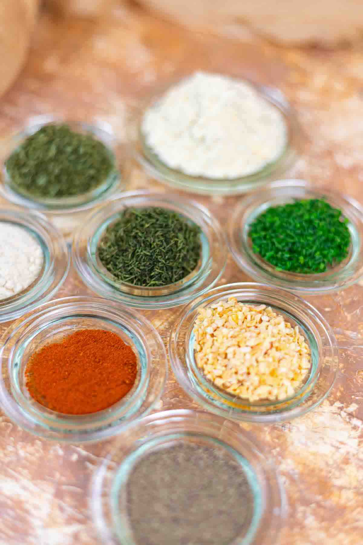 Small glass bowls with assorted dried herbs and spices, including paprika, dried chives, minced onion, dried dill, and parsley, arranged on a light brown surface.
