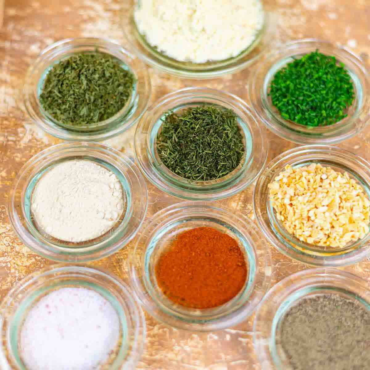 Nine small glass bowls containing various dried herbs and spices, including green herbs, paprika, dried onion, and powders, arranged on a brown textured surface.