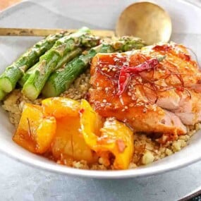 A bowl with grilled chicken, asparagus spears, yellow bell peppers, and quinoa, garnished with sesame seeds and red chili strips.
