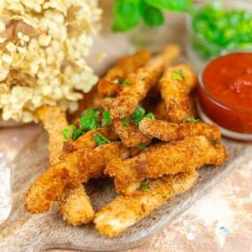 A pile of breaded and fried stick-shaped appetizers on a wooden board, garnished with herbs, served with a bowl of red dipping sauce and a side of chopped green garnish.