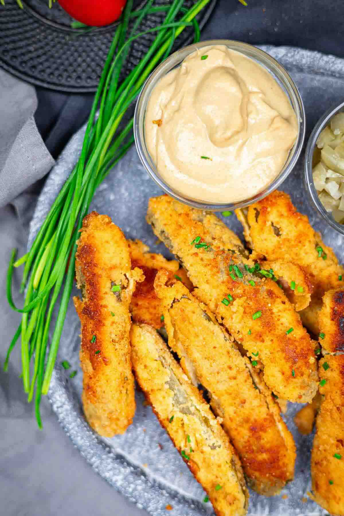 Breaded, fried pickle spears garnished with chopped chives, served with a small bowl of dipping sauce and chopped pickles on a gray plate.