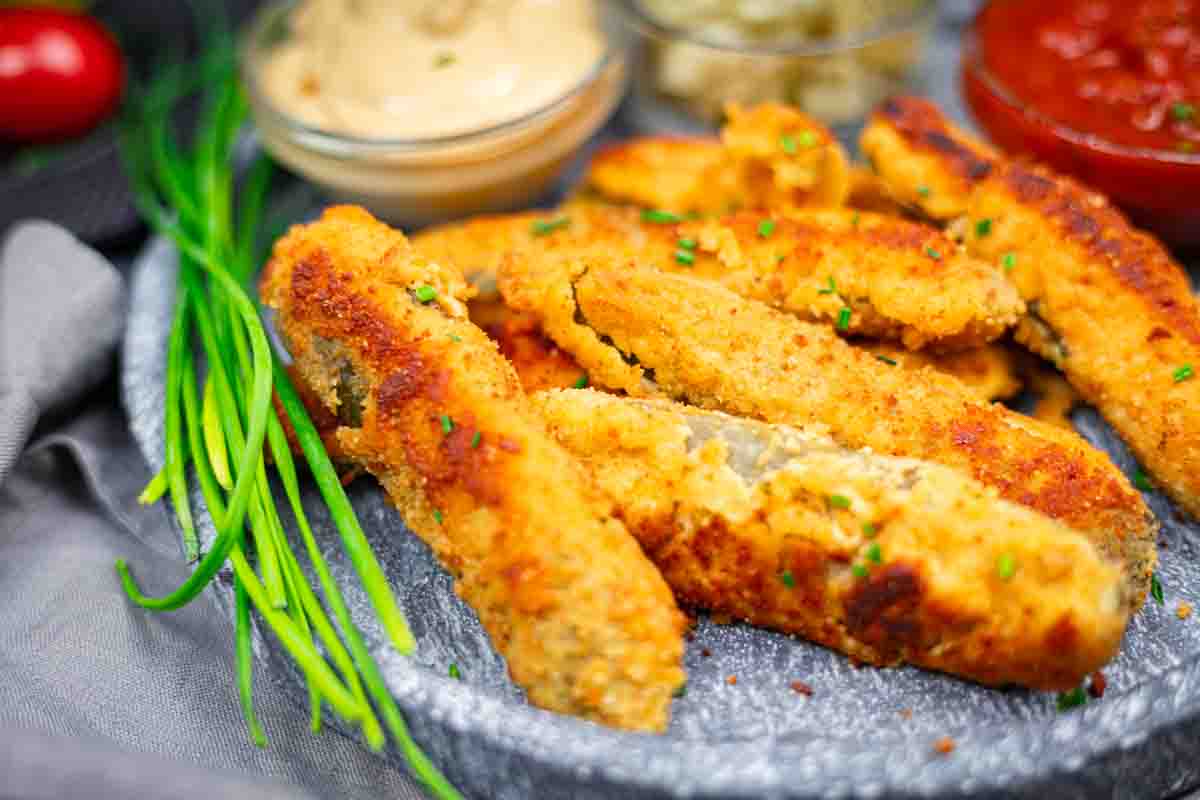 A plate of breaded and fried pickle strips garnished with chives, with dipping sauces in small bowls nearby.