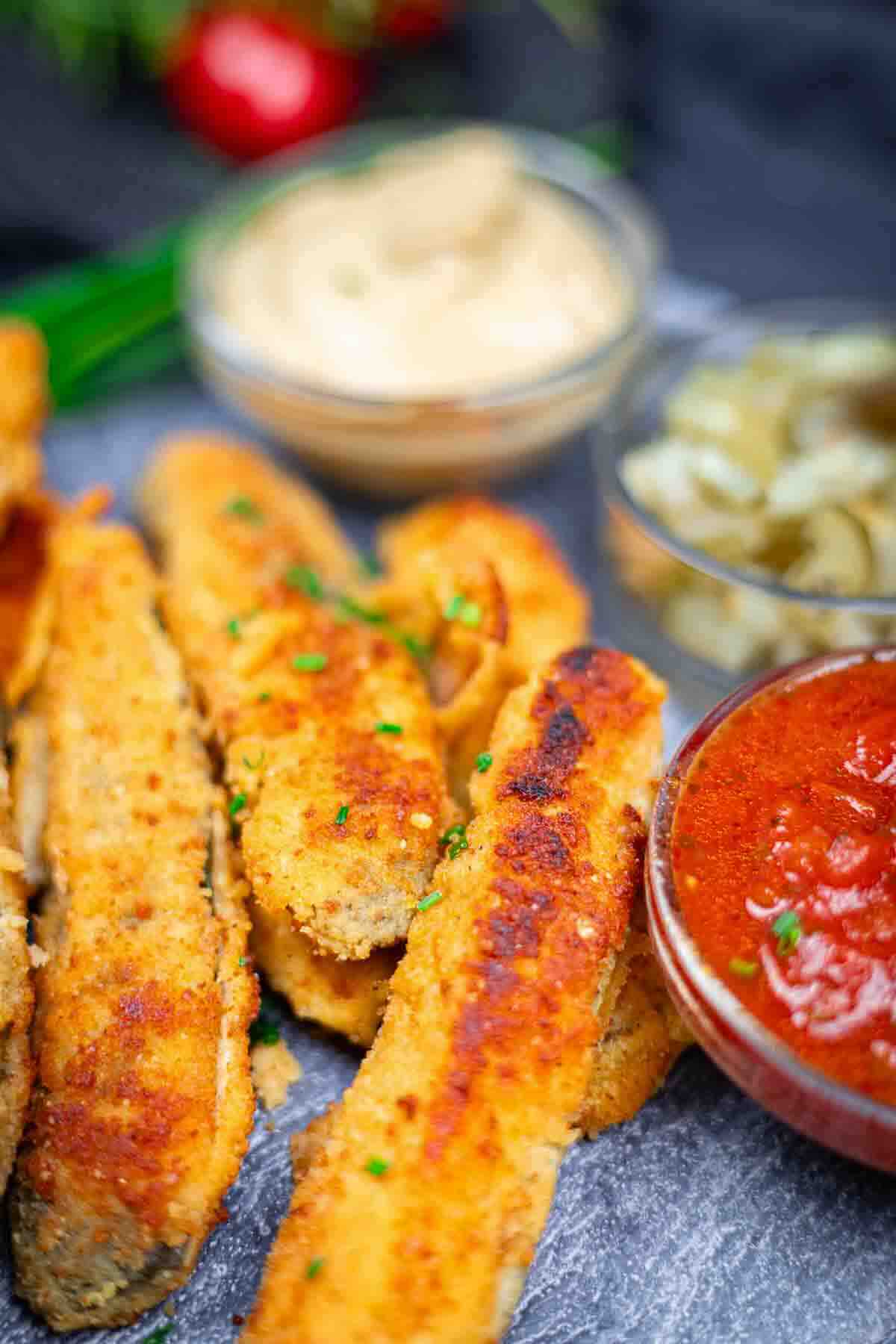 Breaded and fried eggplant sticks served with bowls of creamy dip, pickles, and marinara sauce on a dark surface.