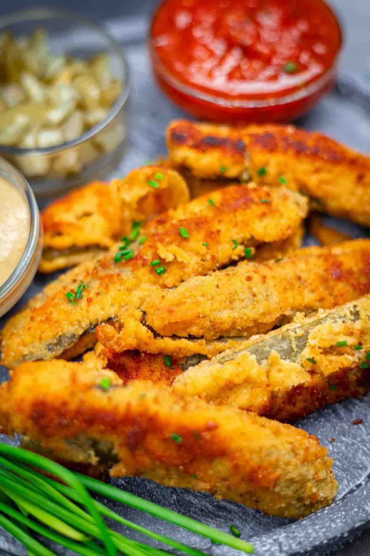 Plate of breaded and fried eggplant strips garnished with chives, served with small bowls of dipping sauces and pickled vegetables.