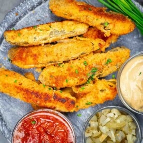 Breaded fried pickle spears arranged on a plate with sides of marinara sauce, chopped onions, and a creamy dipping sauce.