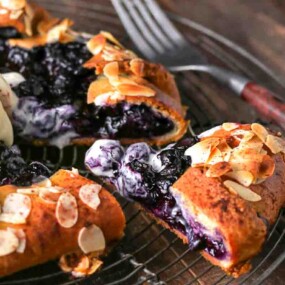 A sliced blueberry galette topped with flaked almonds and a scoop of vanilla ice cream, served on a cooling rack with a fork nearby.