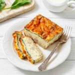 A plate with slices of baked egg casserole topped with melted cheese, accompanied by a fork, on a white wooden table.