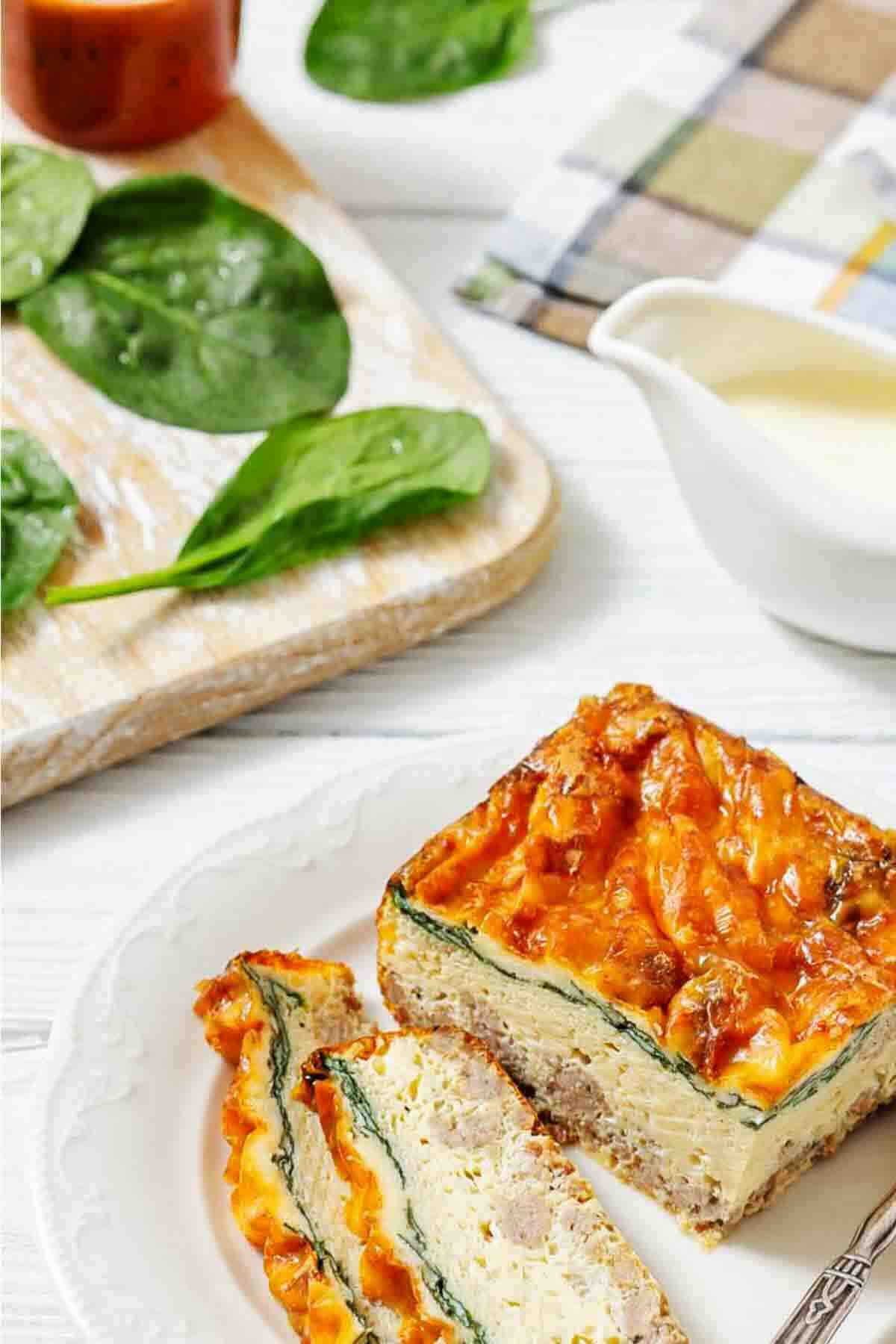 A plate with two slices of egg casserole featuring spinach and melted cheese, next to a cutting board with fresh spinach leaves and a small white sauce pitcher.
