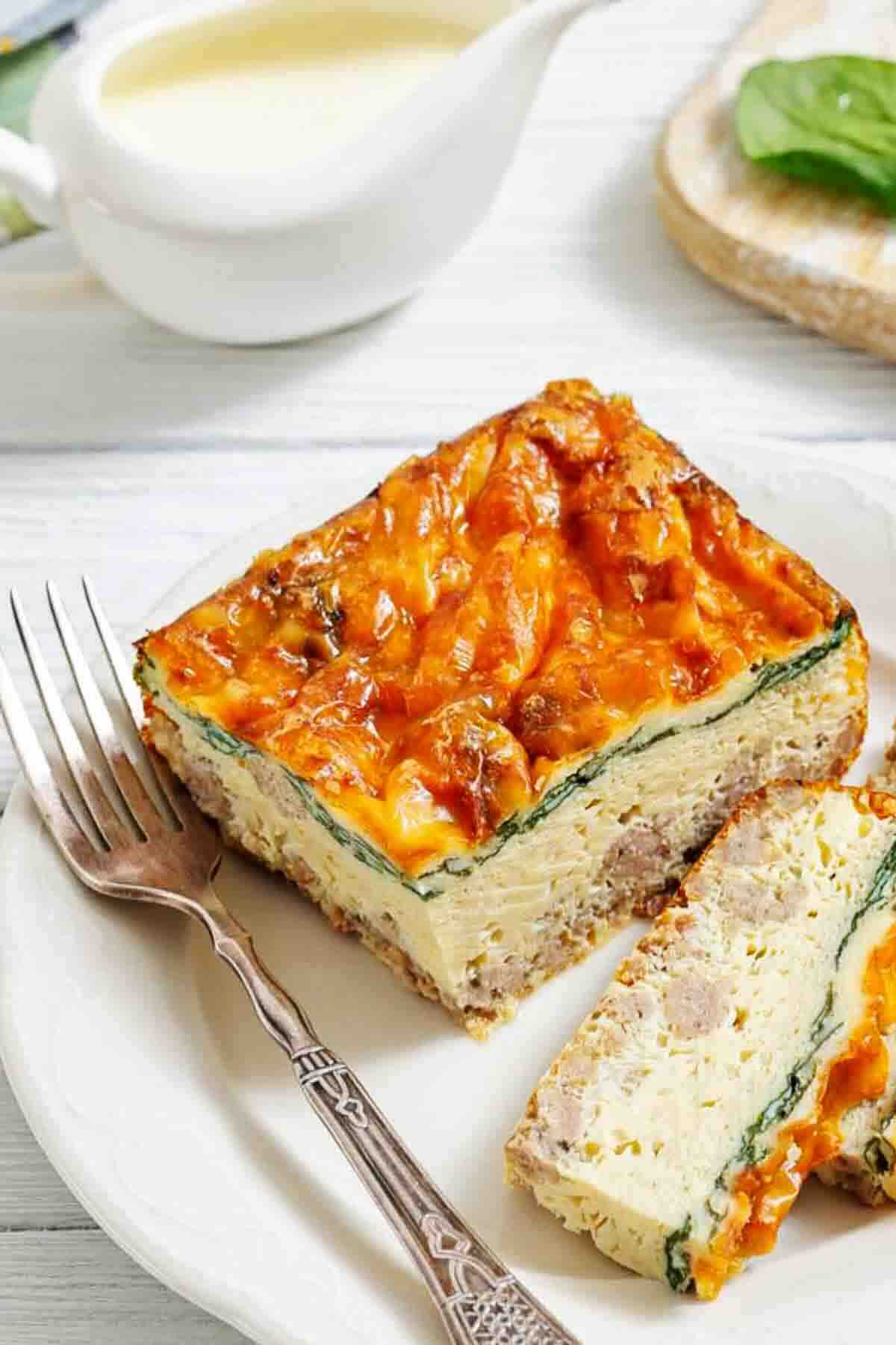 A slice of egg casserole with sausage, cheese, and spinach on a white plate with a fork, next to a small pitcher of sauce.
