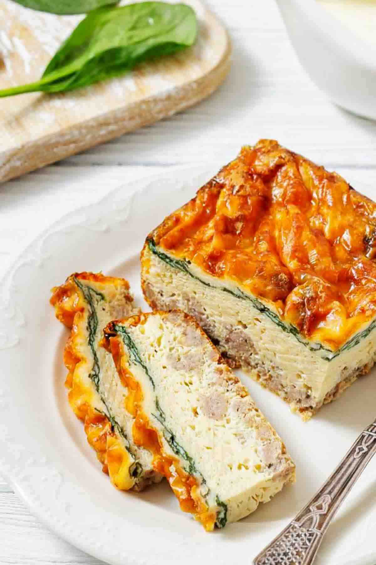 A plate with three slices of baked egg casserole containing sausage, cheese, and spinach, with a fork beside the food.