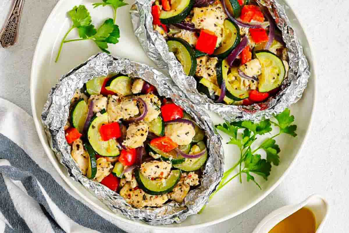 Two foil packets filled with baked chicken, zucchini, red bell pepper, and herbs, served on a white plate with parsley garnish.