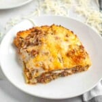 A slice of baked lasagna with a browned cheese topping is served on a white plate, with shredded cheese scattered in the background.