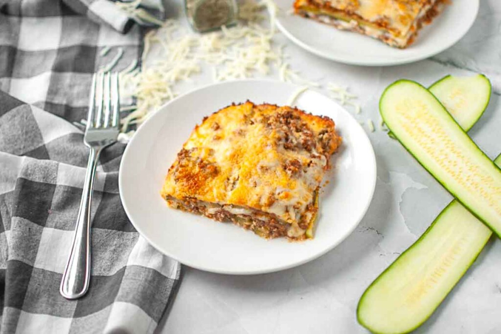 A slice of lasagna on a white plate with a fork, sliced zucchini, shredded cheese, and a checkered napkin nearby.