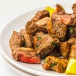 A close-up of cooked steak bites with visible seasoning, garnished with herbs and served on a white plate with pieces of yellow bell pepper.