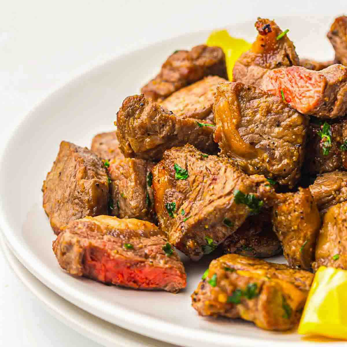 A close-up of cooked steak bites with visible seasoning, garnished with herbs and served on a white plate with pieces of yellow bell pepper.