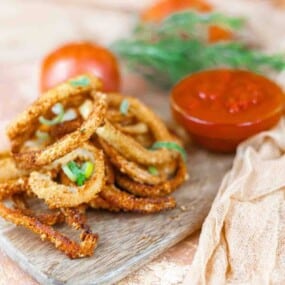 A pile of crispy onion rings garnished with green onions on a wooden board, with a bowl of red dipping sauce and tomatoes in the background.