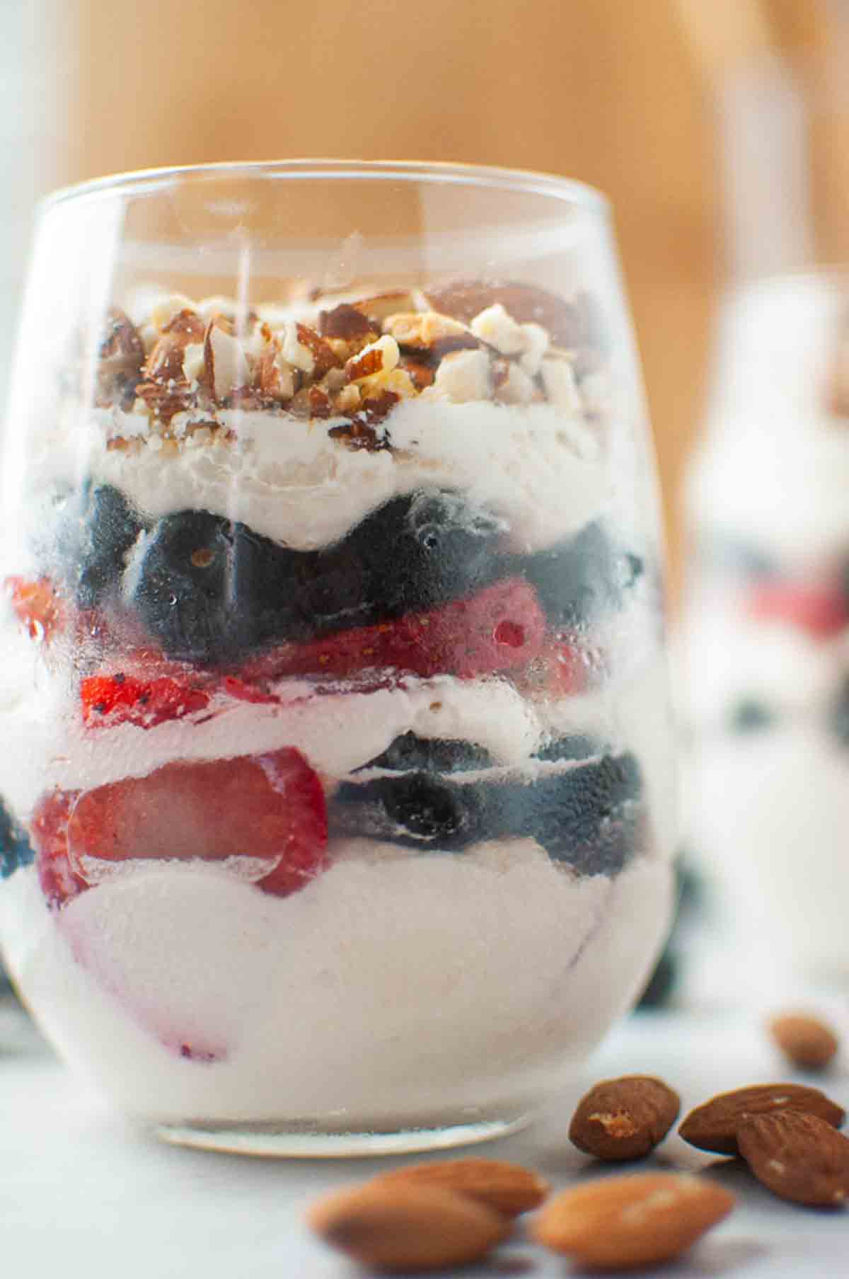 A glass filled with layers of whipped cream, blueberries, strawberries, and chopped nuts, with whole almonds placed beside the glass.