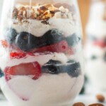Close-up of a layered parfait in a glass, featuring whipped cream, strawberries, blueberries, and chopped nuts.