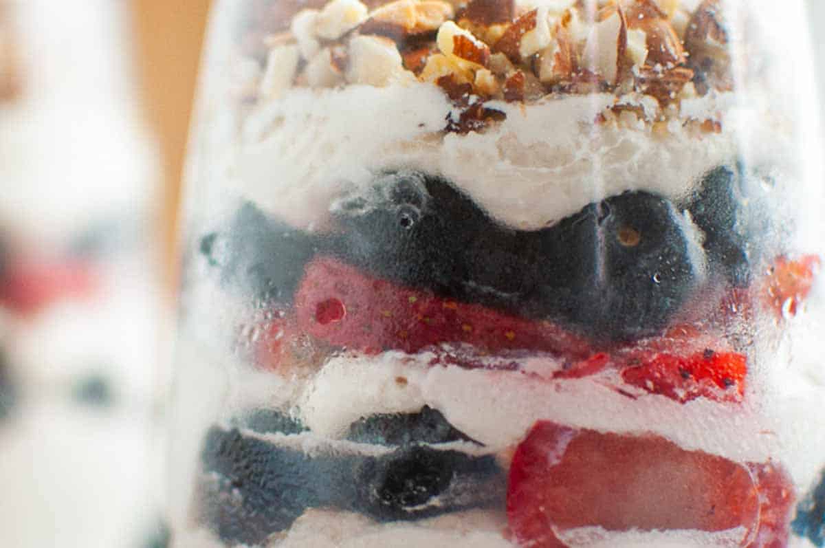 Close-up of a parfait in a glass, showing layers of whipped cream, blueberries, strawberries, and chopped nuts.