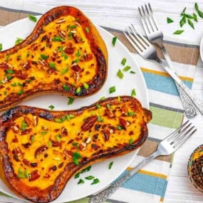 Two halves of roasted butternut squash filled with a creamy mixture, topped with chopped pecans and chives, served on a white plate with forks on a striped tablecloth.