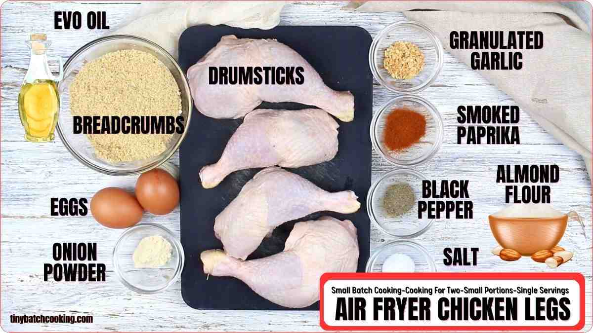 Raw chicken drumsticks on a tray surrounded by bowls of breadcrumbs, almond flour, eggs, spices, and a bottle of oil, labeled for air fryer chicken legs.