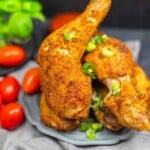 Two pieces of crispy fried chicken garnished with chopped green onions on a gray plate, with cherry tomatoes and basil in the background.