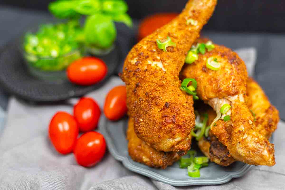 Two crispy fried chicken legs garnished with chopped green onions, served on a plate with cherry tomatoes and green herbs in the background.