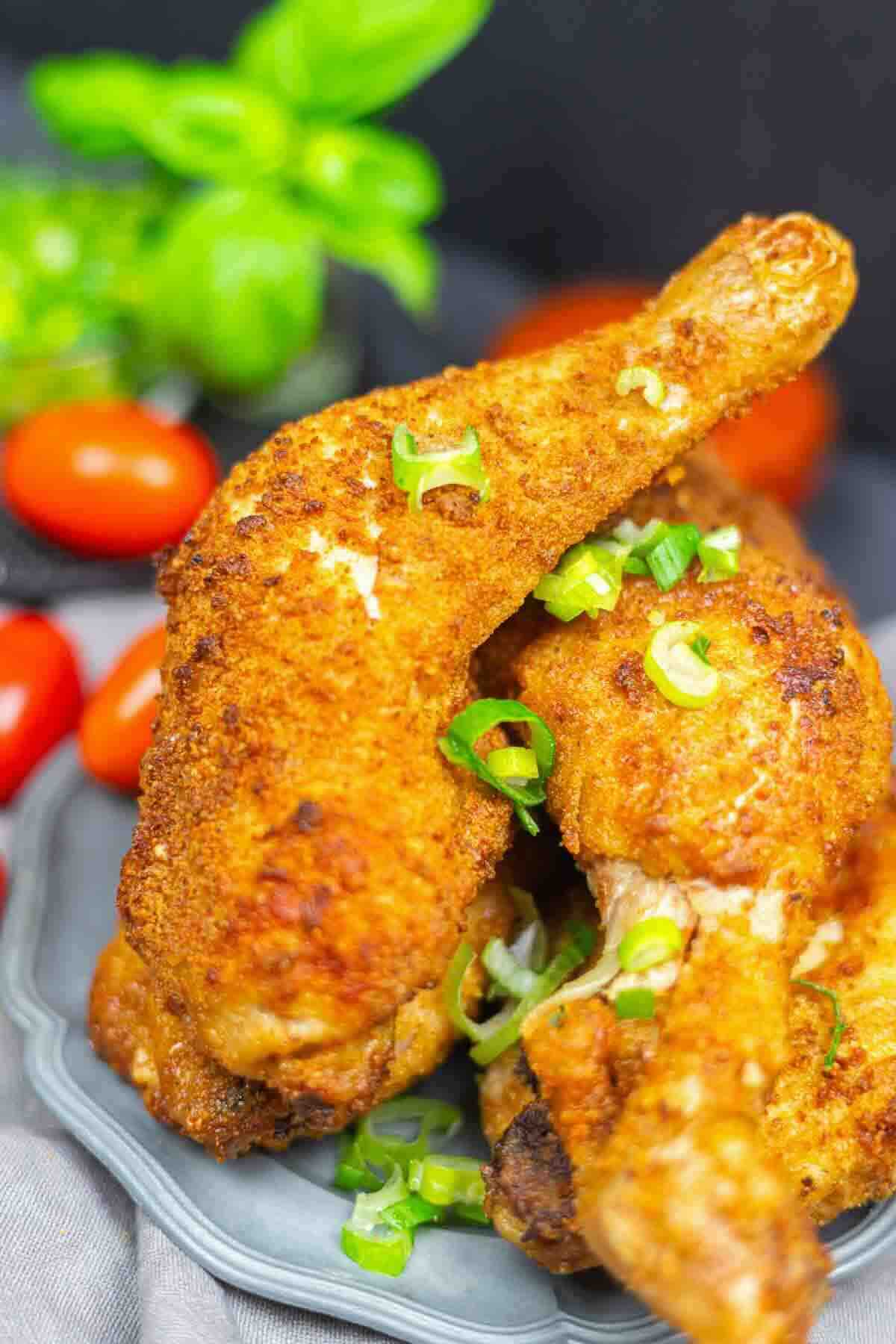 Two pieces of crispy baked chicken legs garnished with chopped green onions on a plate, with tomatoes and fresh basil in the background.