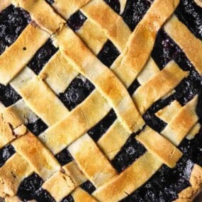 A close-up of a baked pie with a golden brown lattice crust and dark fruit filling, viewed from above.