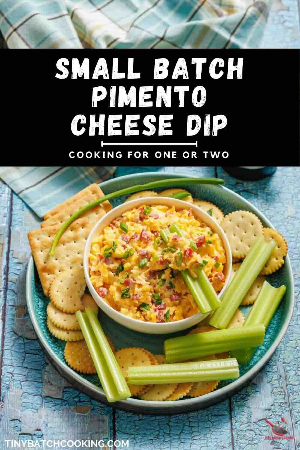 A bowl of pimento cheese dip is surrounded by celery sticks and assorted crackers on a blue plate, set on a plaid cloth.