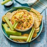 A bowl of pimento cheese dip is surrounded by celery sticks and assorted round and square crackers on a blue plate.