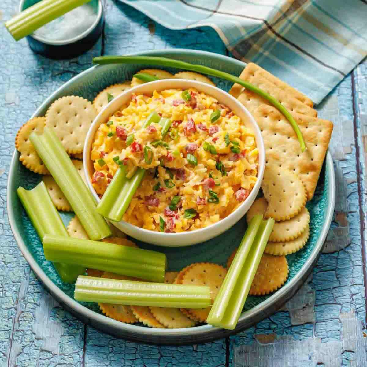 A bowl of pimento cheese dip is surrounded by celery sticks and assorted round and square crackers on a blue plate.