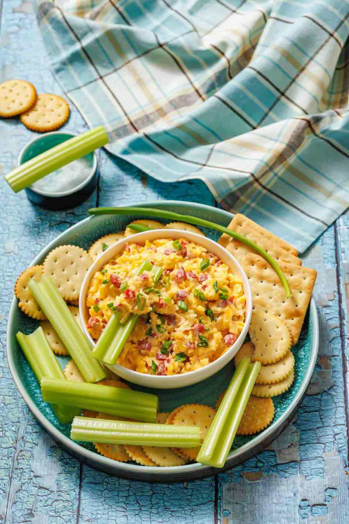 A bowl of cheese and pimento dip surrounded by crackers and celery sticks on a plate, with a plaid cloth and small bowl of dip in the background.