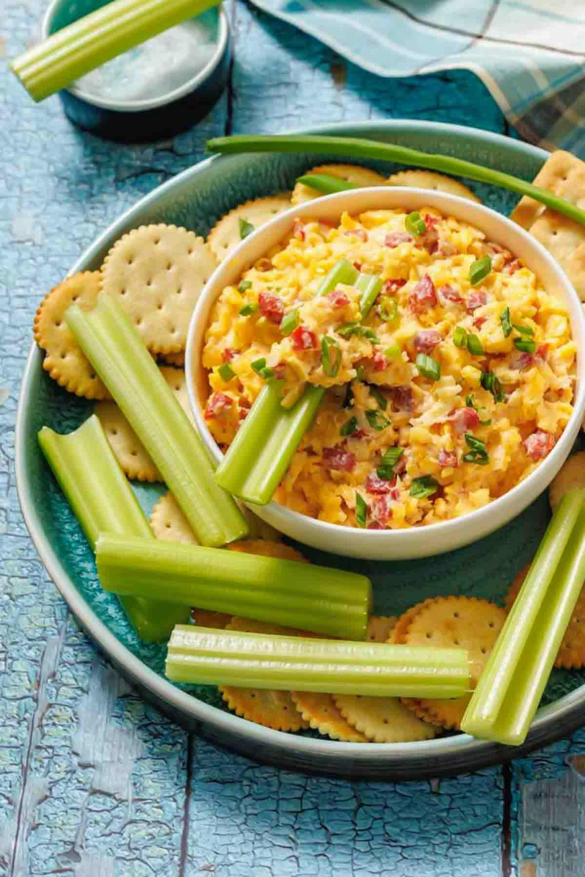 A bowl of pimento cheese dip is served with round crackers and celery sticks on a green plate placed on a cracked blue wooden surface.