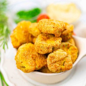 A bowl filled with golden-brown, breaded vegetable nuggets is shown on a white surface, with blurred green herbs and dip in the background.