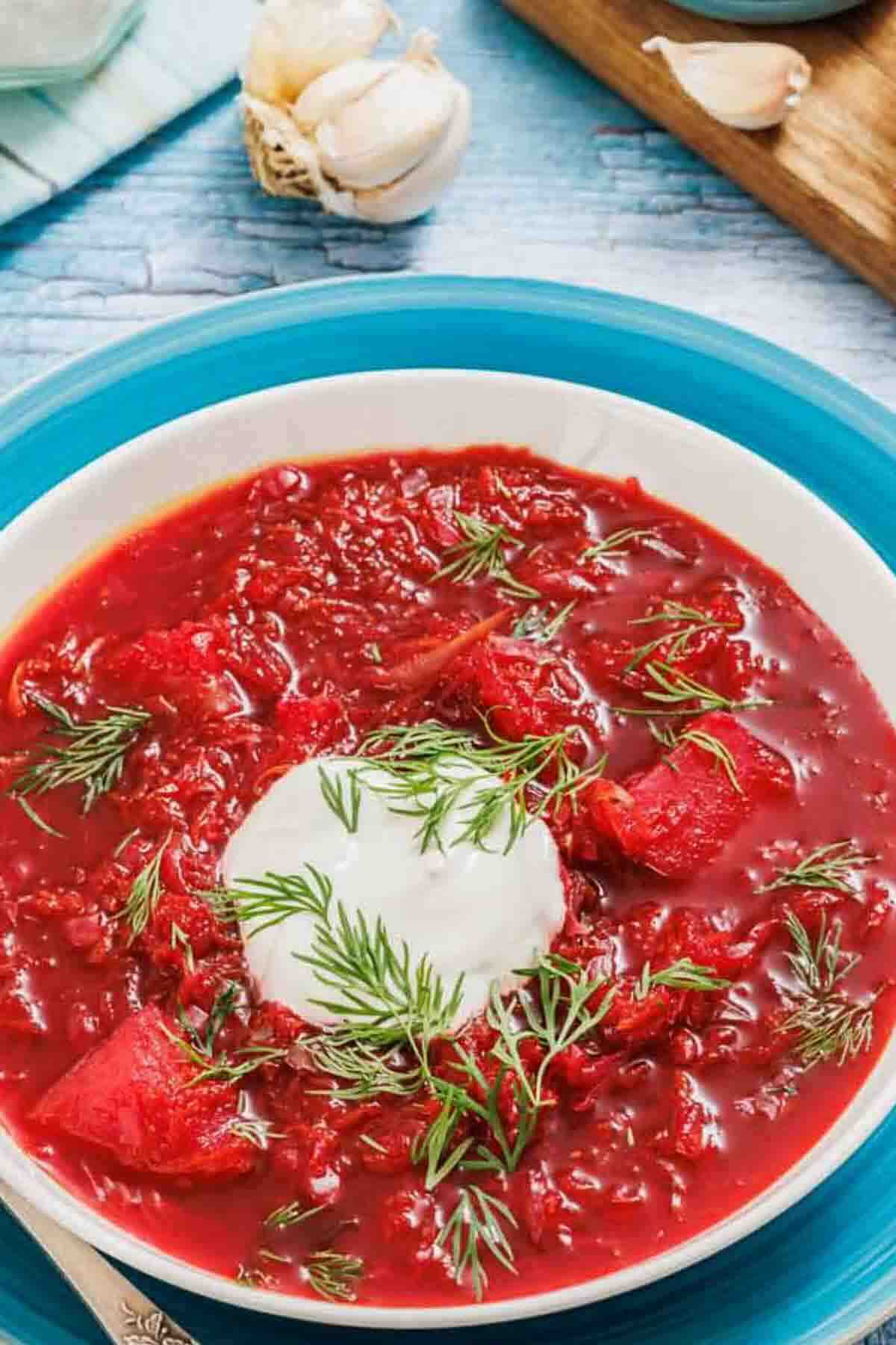 A bowl of red beet soup garnished with a dollop of sour cream and fresh dill, placed on a blue plate with garlic bulbs in the background.