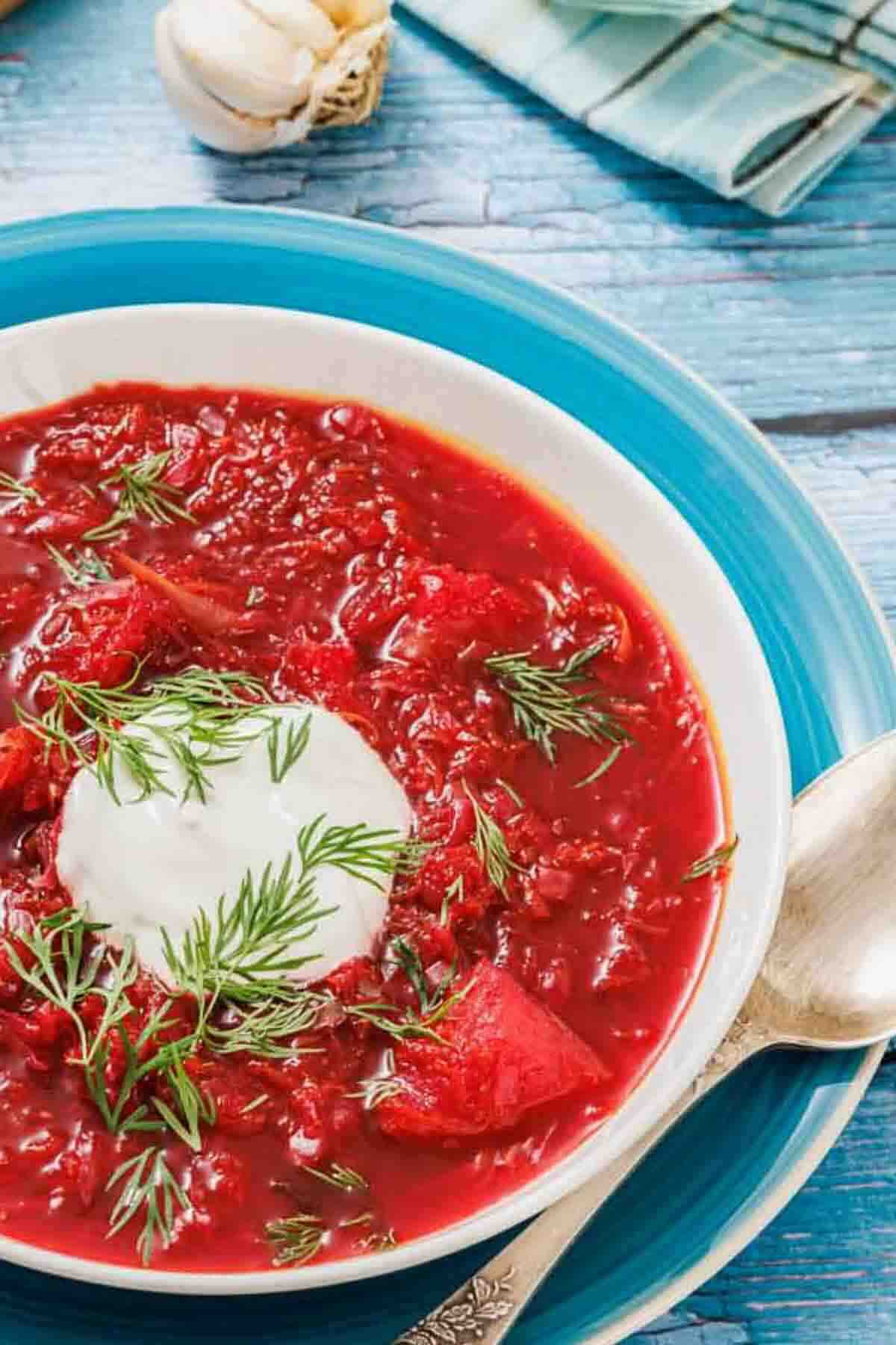 A bowl of borscht topped with a dollop of sour cream and fresh dill, served on a blue plate with a spoon.