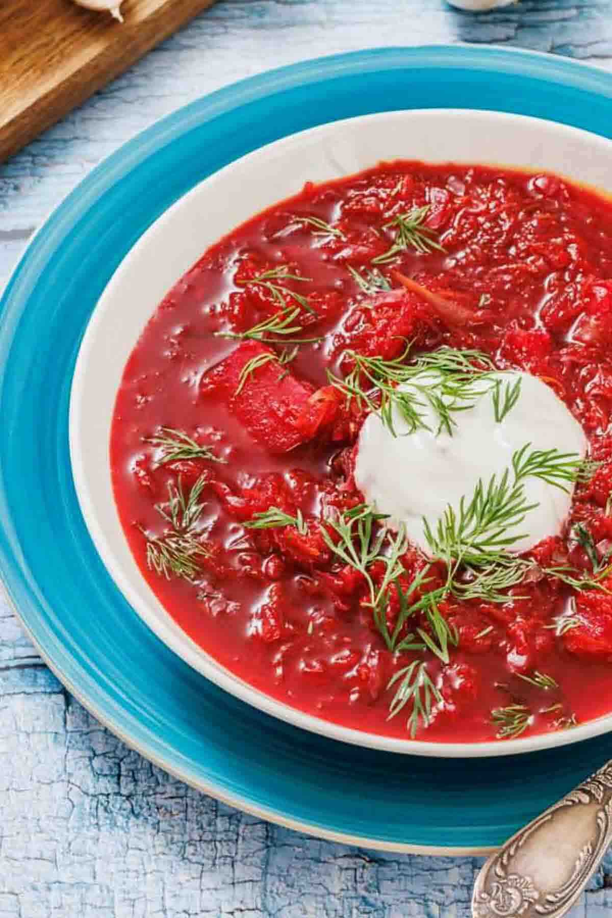 A bowl of red beet soup garnished with a dollop of sour cream and fresh dill, served on a blue plate.