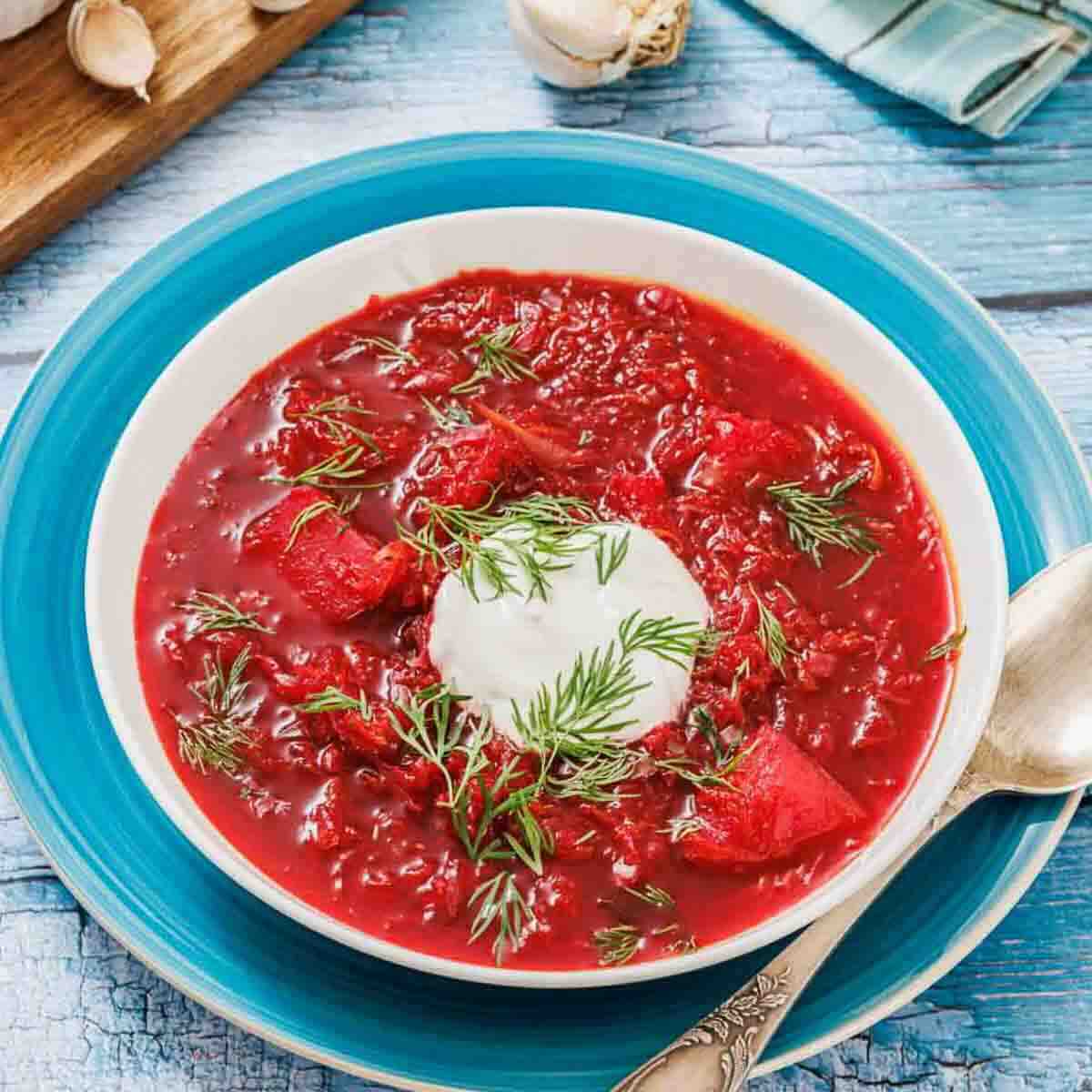 A bowl of borscht soup topped with a dollop of sour cream and fresh dill, placed on a blue plate with a spoon beside it.
