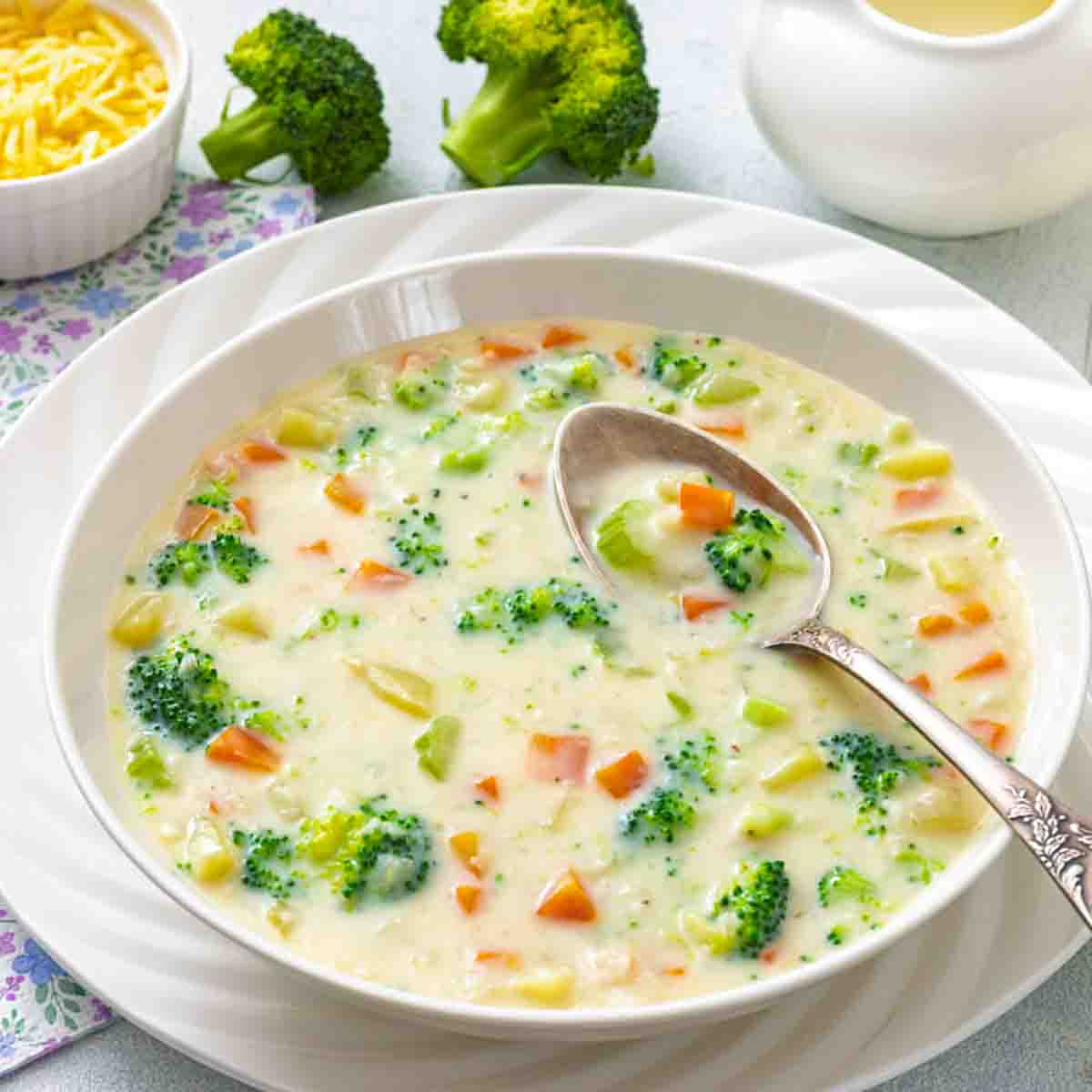 A bowl of creamy broccoli and cheese soup with visible broccoli florets and diced carrots, served with a spoon on a white plate.