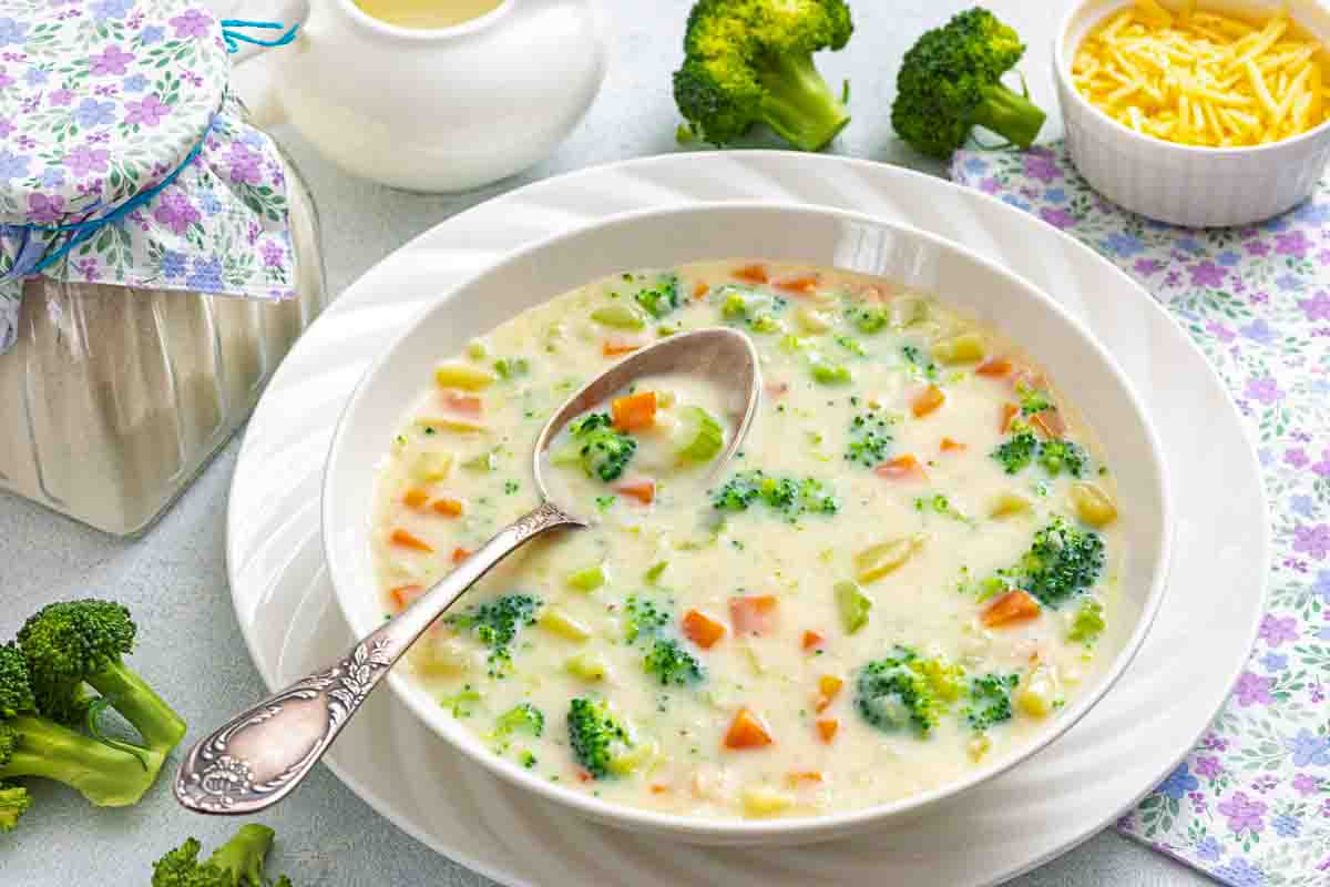 A bowl of creamy broccoli and carrot soup sits on a white plate, with a spoon resting in it. Nearby are pieces of broccoli, shredded cheese, and a floral napkin.