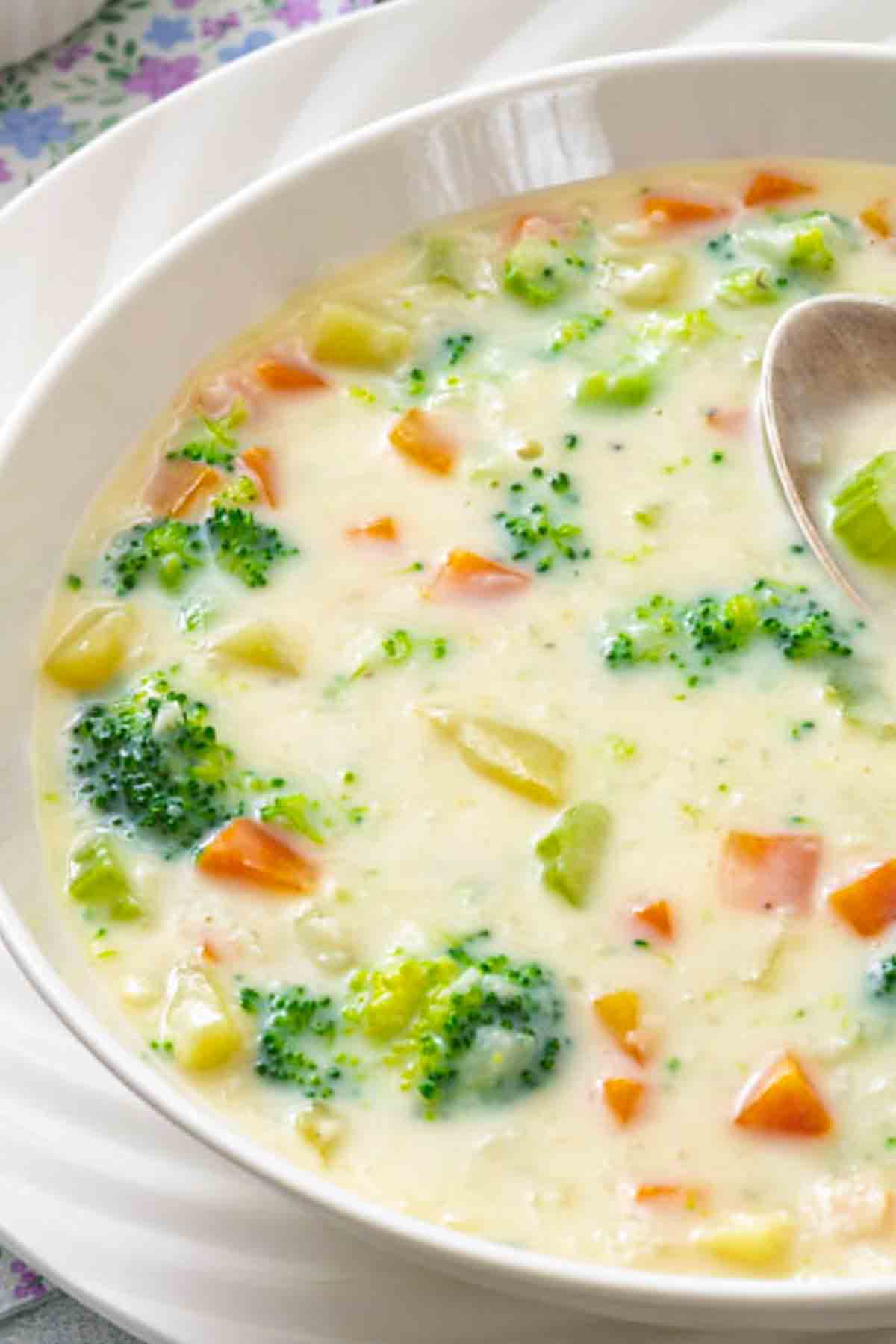 A close-up of a bowl of creamy vegetable soup with visible pieces of broccoli, carrots, and potatoes, served with a spoon.