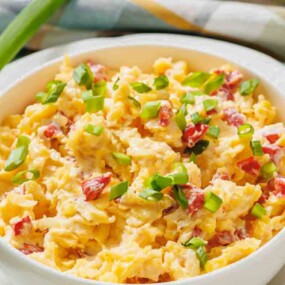 Bowl of scrambled eggs with diced red peppers and chopped green onions on top, placed on a white plate with a green onion beside it.